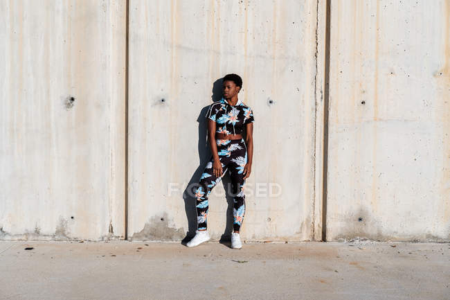 Афроамериканська спортсменка в квітковому спортивному одязі і білі кросівки, дивлячись з цікавістю і сміхом, стоячи на бетонній стіні в сонячних променях і відпочиваючи після тренування в місті. — стокове фото