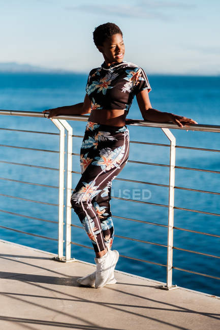Femme afro-américaine en vêtements de sport fleuris et baskets blanches regardant loin avec intérêt et profiter de la vie tout en se tenant appuyé sur la clôture dans les rayons de soleil contre l'eau de mer calme bleu — Photo de stock