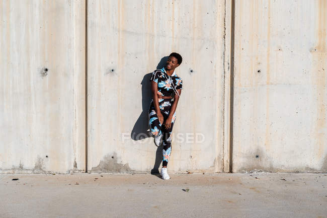 African American female athlete in flowered sport clothes and white sneakers looking away with interest and laughing while standing alone leaning on concrete wall in sunbeams and resting after training in city — Stock Photo