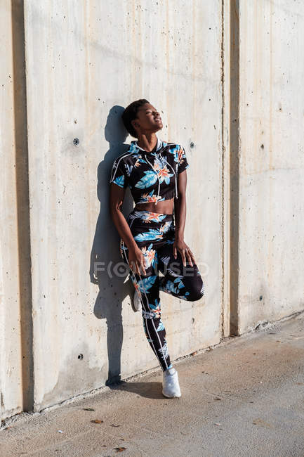 Afrikanisch-amerikanische Sportlerin in geblümter Sportkleidung und weißen Turnschuhen mit geschlossenen Augen, interessiert und lachend, während sie nach dem Training in der Stadt allein an einer Betonwand in Sonnenstrahlen lehnt und ausruht — Stockfoto