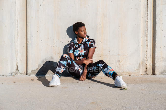 African American female athlete in flowered sport clothes and white sneakers looking away while sitting alone on concrete wall in sunbeams and resting after training in city — Stock Photo