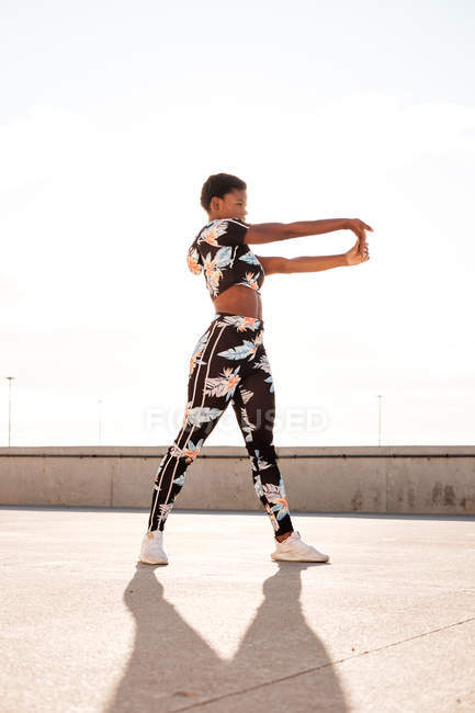 Donna afroamericana adulta in abbigliamento sportivo fiorito che allunga i muscoli della mano mentre sta in piedi da sola e si scalda prima dell'allenamento tra l'ambiente urbano nella giornata di sole — Foto stock