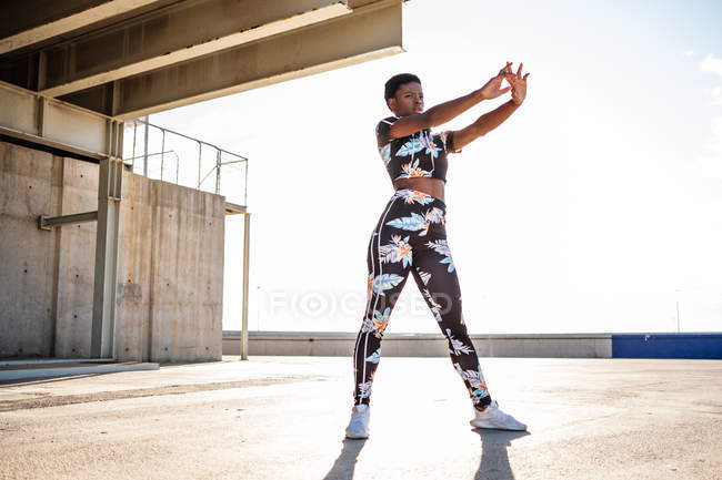 Afrikanisch-amerikanische erwachsene Frau in geblümter Sportbekleidung, die die Handmuskeln dehnt, während sie allein steht und sich vor dem Training in der städtischen Umgebung bei sonnigem Tag aufwärmt — Stockfoto