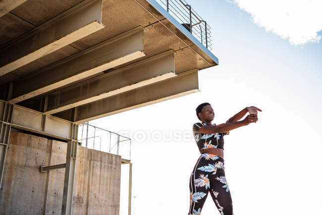 Mujer adulta afroamericana en ropa deportiva con flores que estira los músculos de la mano mientras está sola y se calienta antes de entrenar entre el entorno urbano en un día soleado - foto de stock