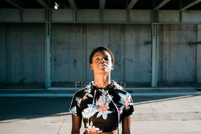 Афроамериканська спортсменка в квітковому спортивному лаві з закритими очима, стоячи на вулиці в сонячних променях до бетонної стіни в місті. — стокове фото