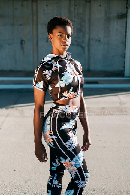African American female athlete in flowered sportswear looking at camera with challenge while standing alone on street in sunbeams against concrete wall in city — Stock Photo