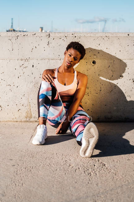 Atleta afroamericana con ropa deportiva florecida y zapatillas blancas mirando a la cámara mientras está sentada sola en la pared de hormigón en rayos de sol y descansando después de entrenar en la ciudad - foto de stock