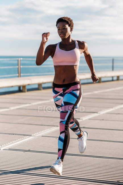 African American adult sportswoman in vibrant green activewear focusing and running alone along waterfront among metal columns under roof — Stock Photo