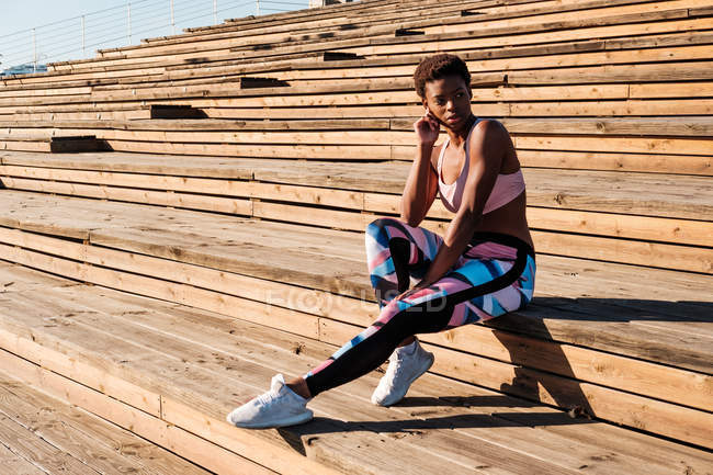 Mujer joven afroamericana en ropa deportiva colorida y zapatillas blancas mirando hacia otro lado con curiosidad y contemplando mientras está sentada sola en escaleras de madera marrón en el estadio - foto de stock