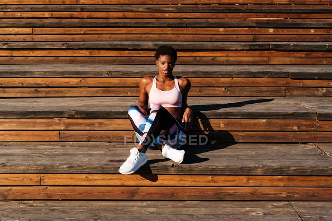Молодая афроамериканка в разноцветной спортивной одежде и белых кроссовках с любопытством смотрит в камеру и размышляет, сидя в одиночестве на коричневой деревянной лестнице на стадионе — стоковое фото