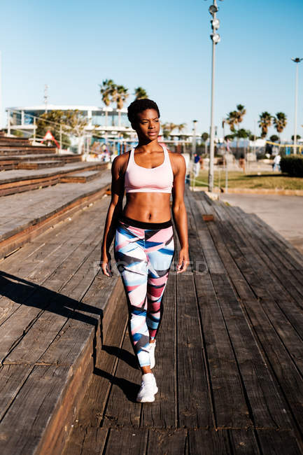Афроамериканская девушка в разноцветной спортивной одежде и белых кроссовках с любопытством смотрит в сторону и созерцает прогулку в одиночестве по коричневой деревянной лестнице на стадионе — стоковое фото