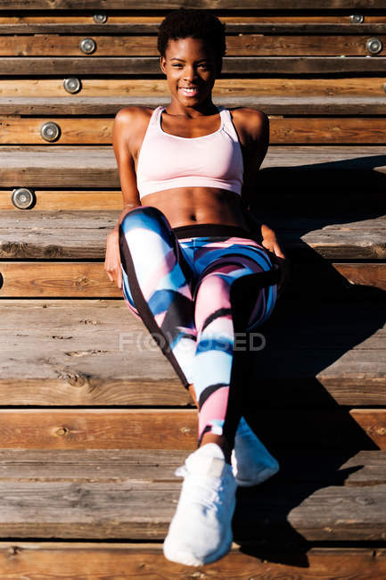 Mujer joven afroamericana en ropa deportiva colorida y zapatillas blancas mirando a la cámara con curiosidad y contemplando mientras está sentada sola en escaleras de madera marrón en el estadio - foto de stock