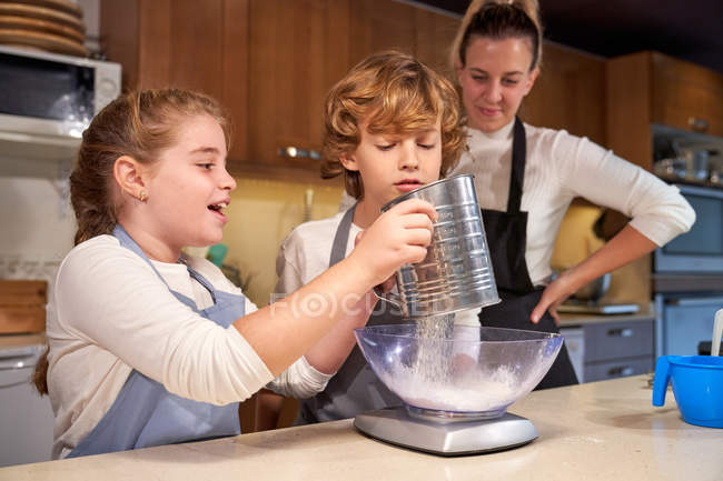 Girl pouring sugar into bowl with boy and woman in kitchen — Stock Photo