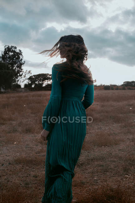 Visão traseira da fêmea irreconhecível em vestido elegante em pé no prado à noite ventosa e nublada na natureza — Fotografia de Stock