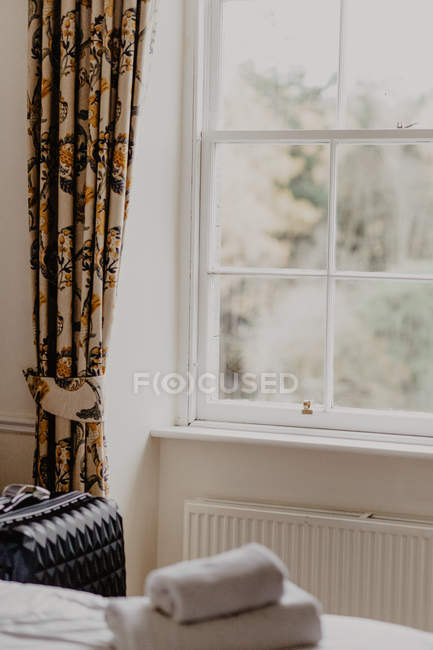 Valise placée près de la fenêtre décorée d'un rideau floral dans une chambre d'hôtel lumineuse — Photo de stock