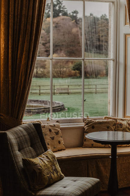 Стіл розташований поруч з м'яким кріслом і диваном з подушками проти вікна з видом на сад в осінній день — стокове фото