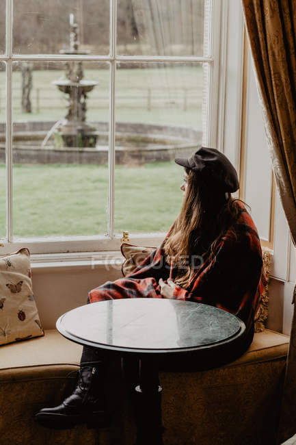 Вид сбоку молодой женщины, завернутой в теплое одеяло, сидящей за столом на диване и выглядывающей из окна в уютной комнате рядом с садом — стоковое фото