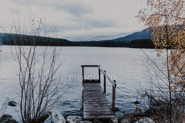 Wide river with wooden pier near forest in fall on cloudy daytime — Stock Photo