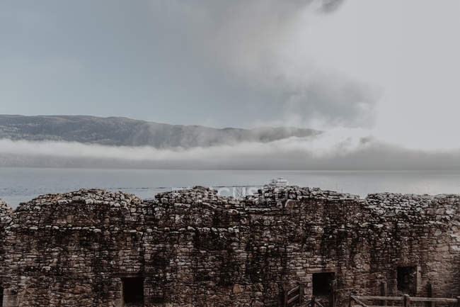 Pared de piedra del viejo castillo en ruinas contra el cielo nublado con vista a las montañas brumosas - foto de stock