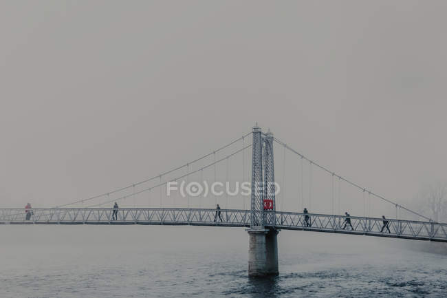 Люди, що йдуть через річку на сучасному мосту з туманом в похмурий день — стокове фото