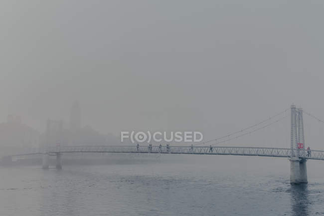 Les gens marchent à travers la rivière sur un pont moderne avec de la brume pendant la journée nuageuse — Photo de stock