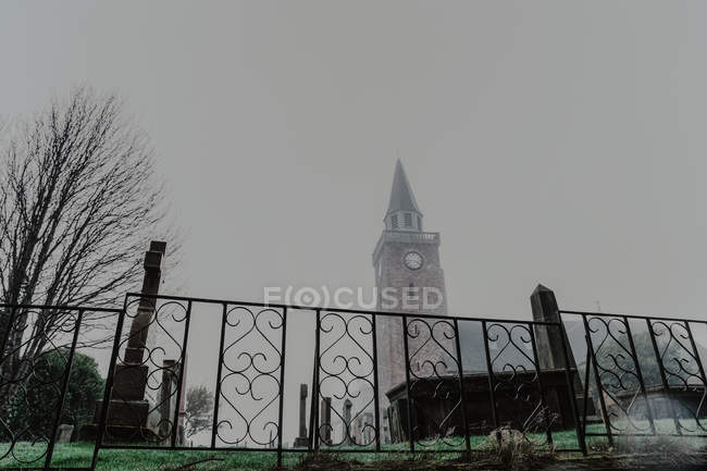 From below of foggy graveyard with fence and aged church on cloudy daytime — Stock Photo