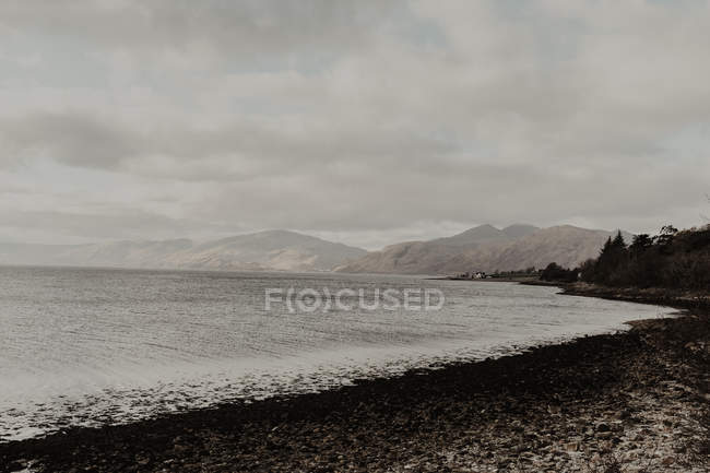 Leeres welliges Wasser spült dunkles Ufer umgeben von Bergen unter grauem bewölkten Himmel — Stockfoto
