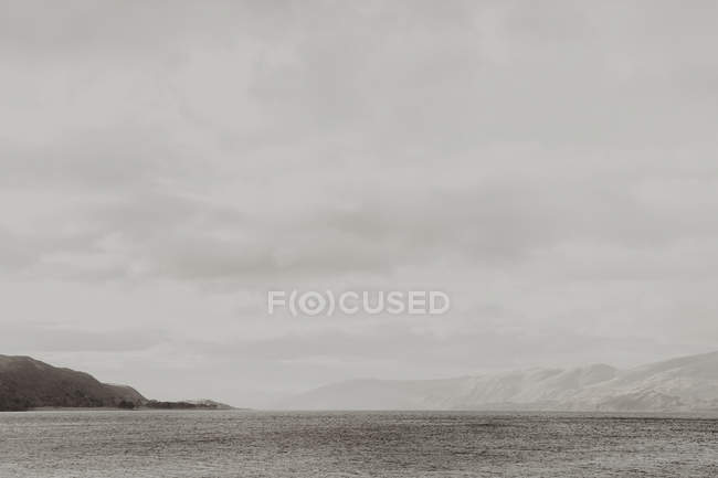 Leeres welliges Wasser spült dunkles Ufer umgeben von Bergen unter grauem bewölkten Himmel — Stockfoto