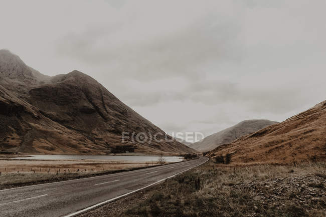 Route étroite et balisée longeant une rivière calme au pied de montagnes pierreuses sous un ciel gris — Photo de stock
