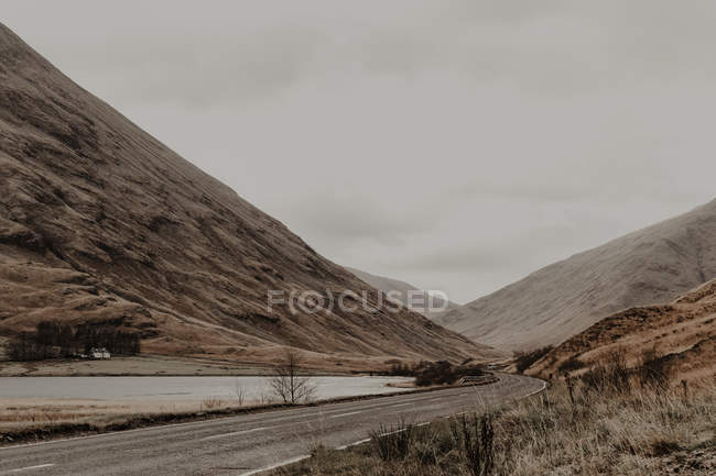 Schmale markierte Straße entlang des ruhigen Flusses am Fuße steinerner Berge unter grauem Himmel — Stockfoto