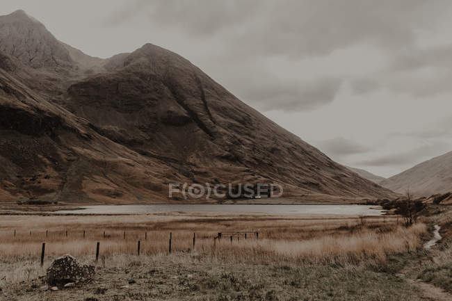 Narrow marked road going along calm river at foot of stony mountains under gray sky — Stock Photo