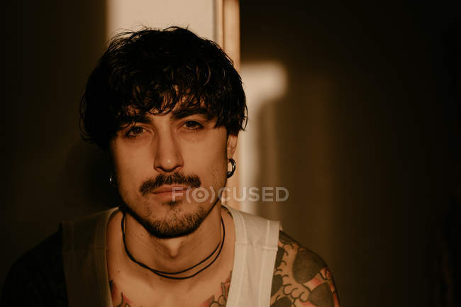 Joven hombre hipster con estilo con tatuajes en camiseta blanca apoyada contra la pared, mirando en la cámara - foto de stock