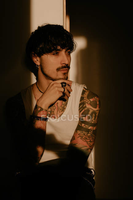 Jeune homme avec moustache et tatouages en débardeur blanc debout avec la main sur le menton — Photo de stock