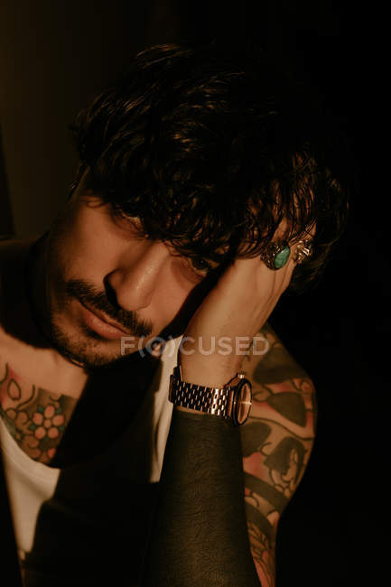 Молодой красивый мужчина с усами и татуировками, опирающийся на руку и смотрящий в камеру — стоковое фото