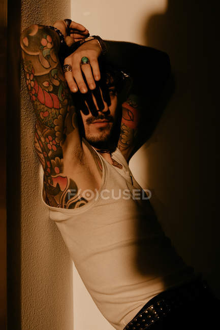 Jovem homem bonito com bigode e tatuagens encostadas à parede na sombra — Fotografia de Stock