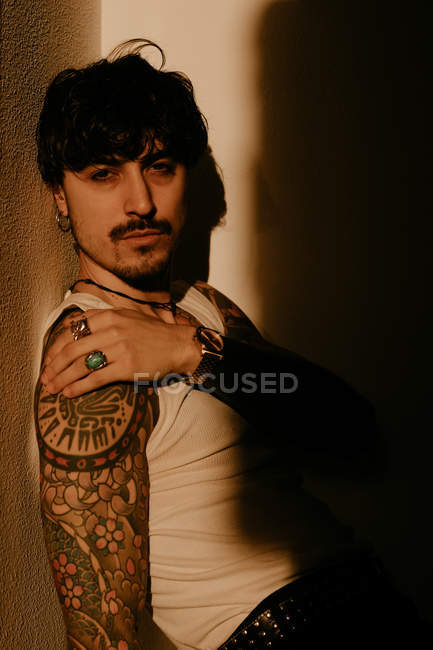 Молодой красивый мужчина с усами и татуировками, прислонившийся к стене в тени — стоковое фото