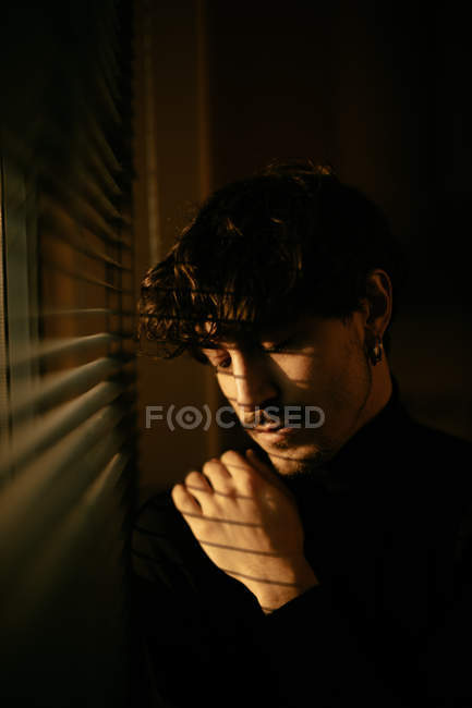 Молодой меланхолик в черной водолазке стоит у окна с жалюзи с тенью на лице — стоковое фото