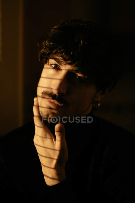 Young melancholic guy in black turtleneck standing next to window with shutters with shadow on face looking in camera — Stock Photo