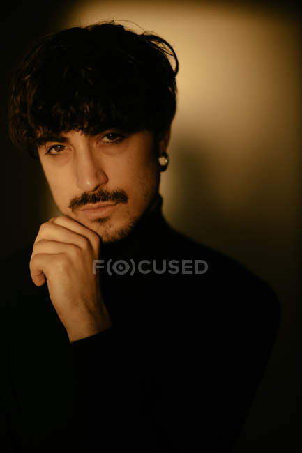 Jeune homme avec moustache à côté du mur regardant dans la caméra avec la main sur le menton — Photo de stock