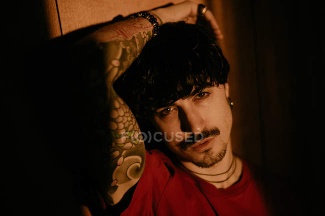 Junger Mann mit Schnurrbart sitzt mit erhobenem Arm an der Wand und blickt in die Kamera — Stockfoto