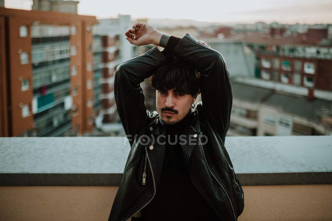 Homem sério em jaqueta de couro em pé no telhado com braços levantados e olhando com confiança na câmera — Fotografia de Stock