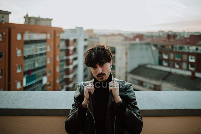 Homem sério em jaqueta de couro em pé no telhado e segurando colarinho de casaco — Fotografia de Stock