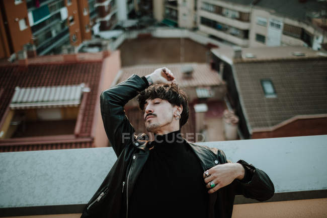Серьезный человек в кожаной куртке, стоящий на крыше и опирающийся на ограждение с закрытыми глазами — стоковое фото