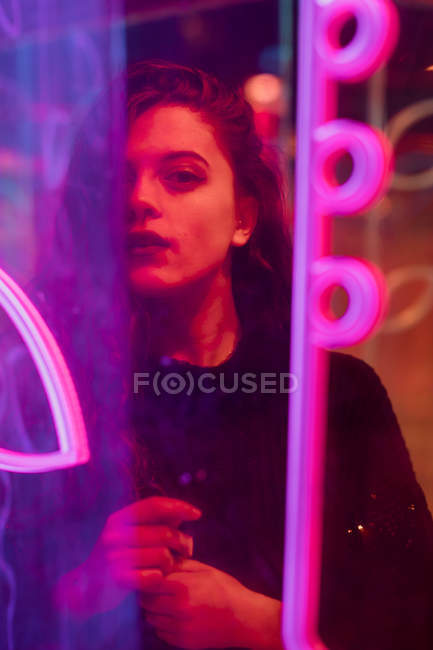 Stylish long-haired brunette in casual wear looking in camera among neon signs at city street — Stock Photo