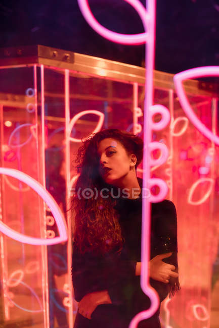Молодая длинноволосая женщина в модной одежде, смотрящая в камеру среди неоновых знаков на городской улице — стоковое фото