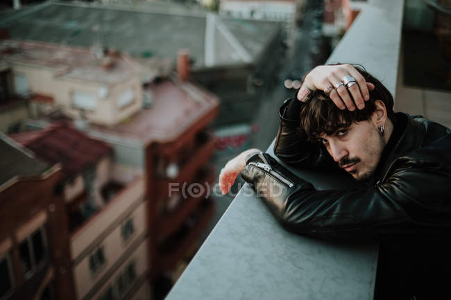 Homem sério em jaqueta de couro apoiando-se na esgrima do telhado e olhando com confiança na câmera — Fotografia de Stock