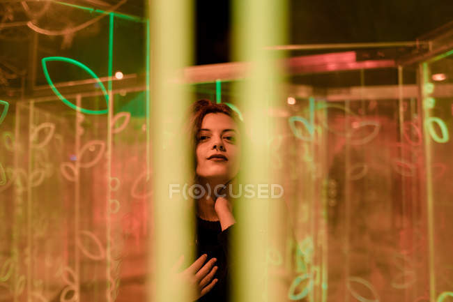 Young long-haired woman in trendy wear looking in camera among neon signs at city street — Stock Photo