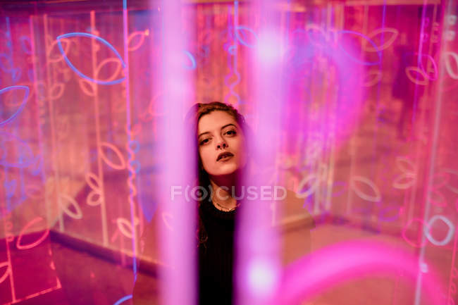 Giovane donna con trucco elegante guardando in macchina fotografica come in piedi tra i segni al neon in strada della città — Foto stock