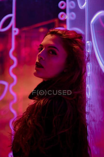 Stylish melancholic brunette in lights of neon signs leaning on wall at city street — Stock Photo