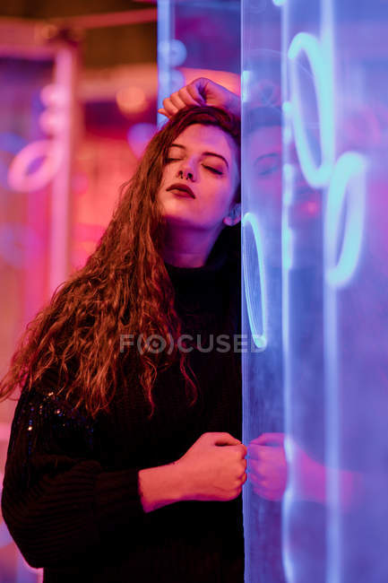 Pretty pensive young woman leaning on wall at city street in neon signs lights — Stock Photo